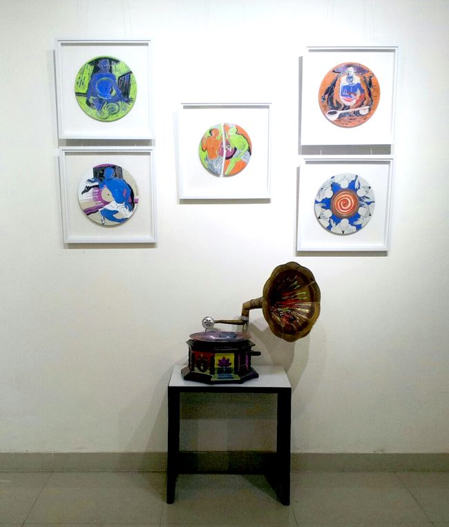 Six painted records and plates in white frames hung above an antique gramophone with a brass horn